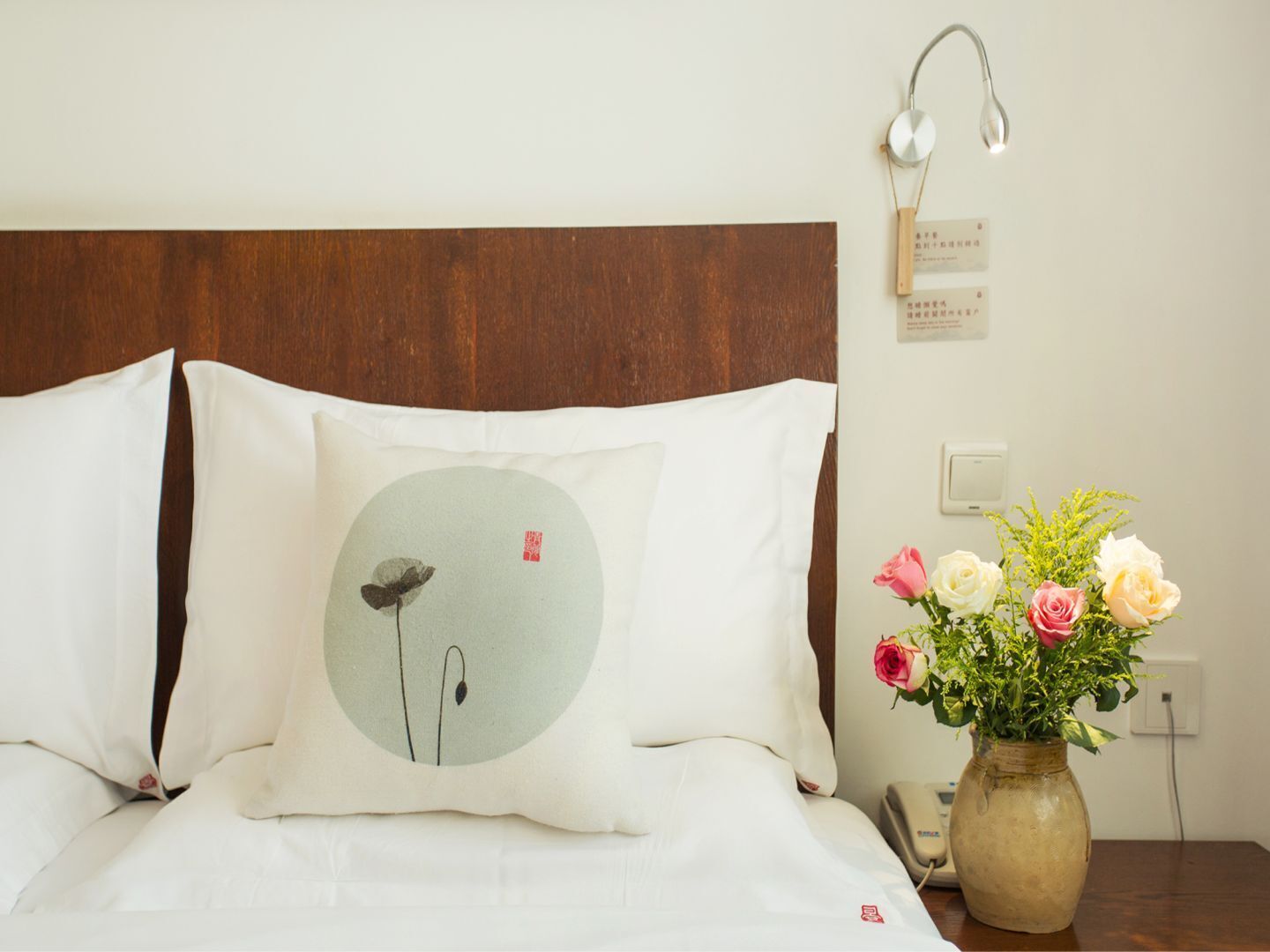 A neatly made bed with white linens and a pillow featuring a minimalist design. A bedside table holds a vase with flowers and a telephone.