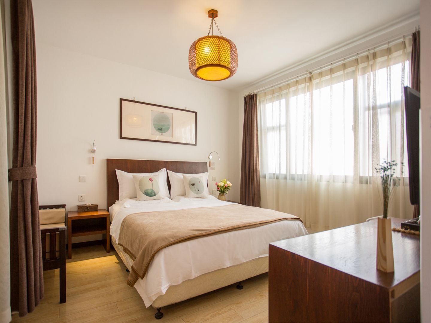 A hotel room with a bed, bedside tables, a desk, a hanging lamp, a framed picture, and a window with curtains.