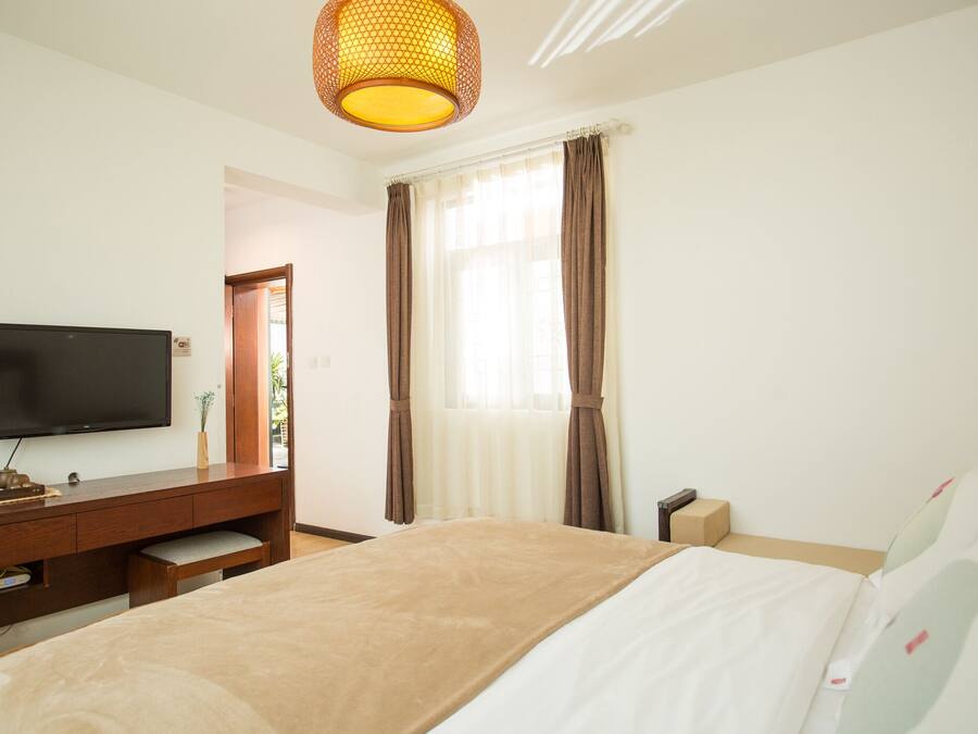 Habitación de hotel con una cama, un televisor sobre una consola de madera, una ventana con cortinas y una lámpara colgante.