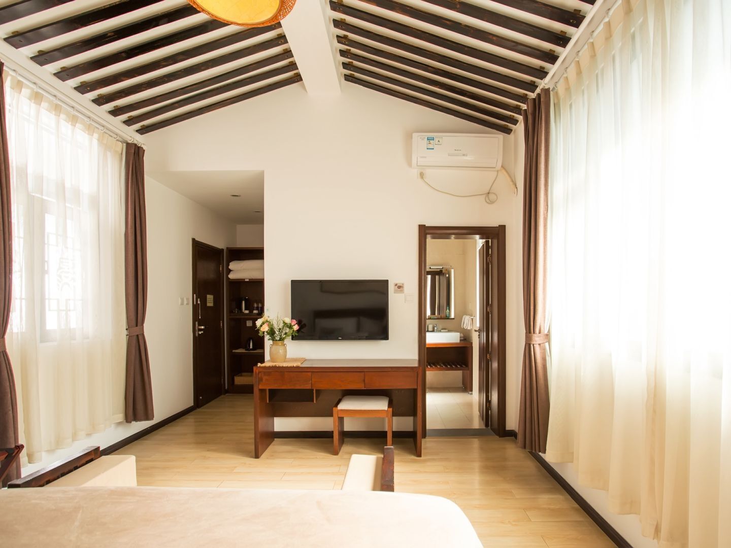 Ein Hotelzimmer mit Holzboden, einem Wand-Fernseher, einem Holztisch mit einer Bank und einem großen Fenster mit Vorhängen.