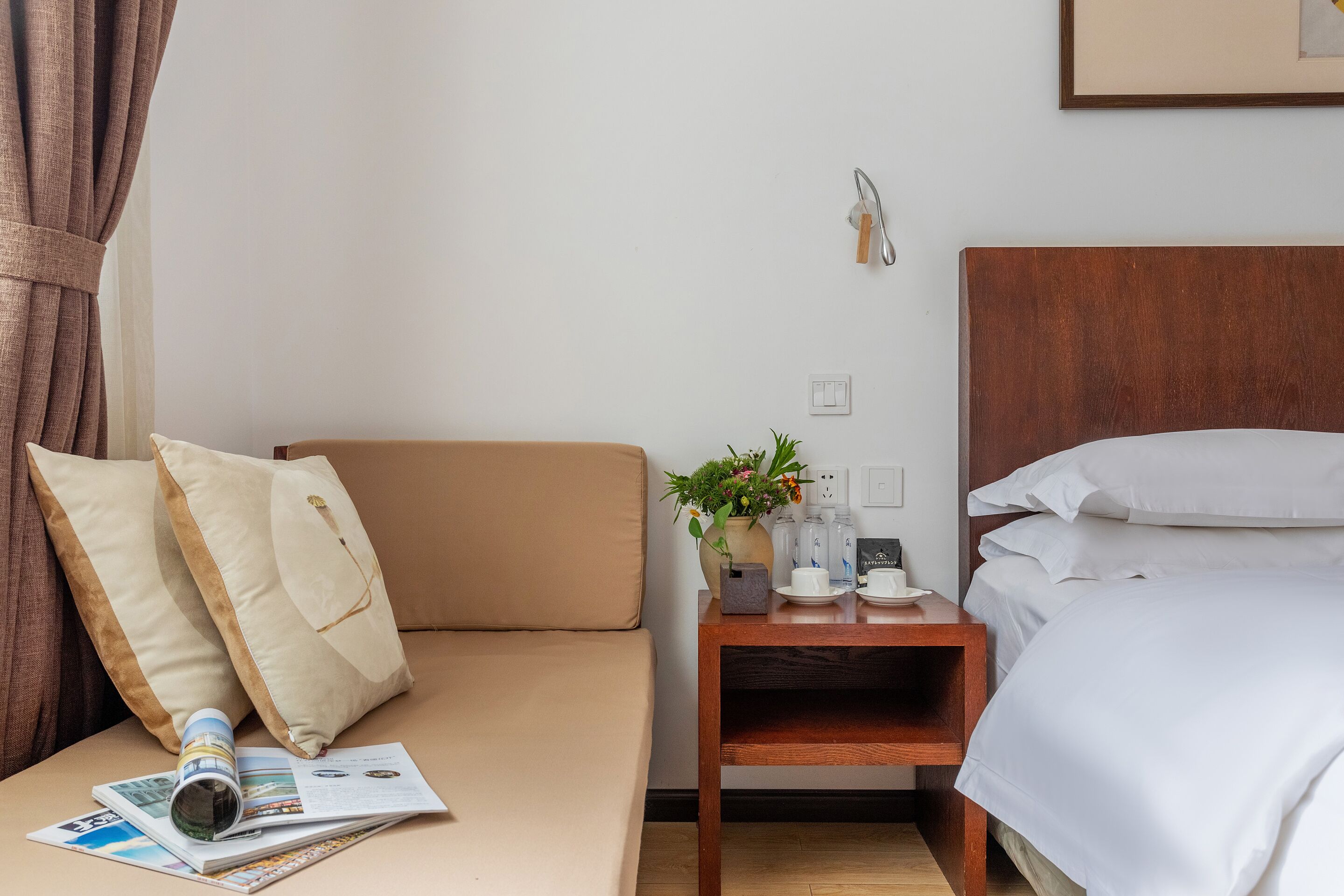 Una habitació d'hotel amb un llit, un sofà, una tauleta de nit de fusta amb un ram de flors, i una revista sobre el sofà.