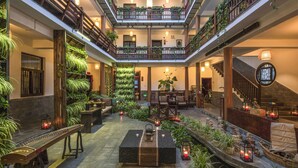Een ruime atriumruimte met een hoge plafond, meerdere balkons met beplanting, een centraal zitgedeelte met tafels en stoelen, en een trap die naar de bovenverdiepingen leidt.