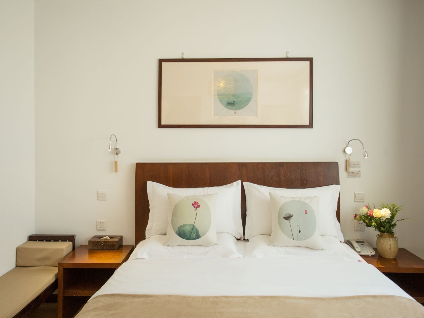 A neatly arranged hotel room with a wooden headboard, two bedside tables with lamps, a small bedside table with a vase of flowers, and a framed picture above the bed.