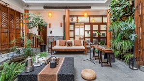 Een ruime, overdekte ruimte met een houten bank, een bankstel en diverse planten. Er zijn houten deuren en een balkon met een hekwerk.