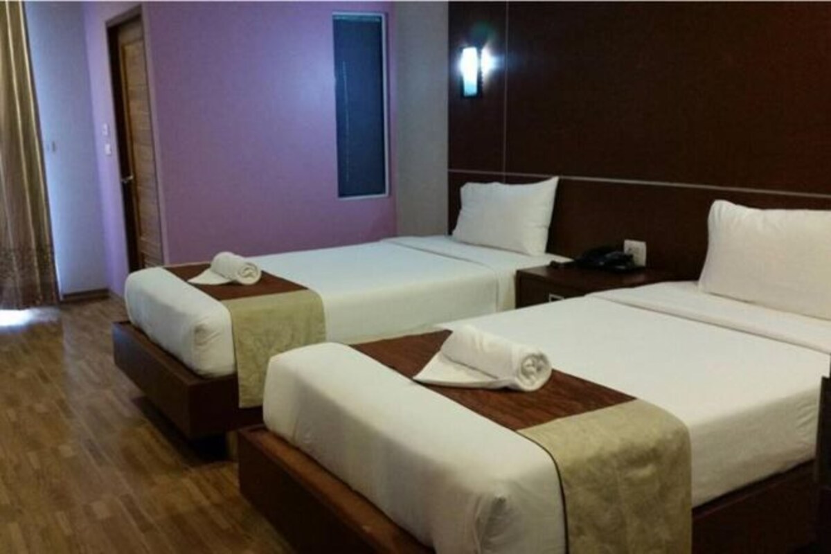 A hotel room with two single beds, a wooden headboard, a small bedside table with a lamp, and a window with blinds.