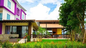 Un edificio moderno con fachada morada y blanca, un espacio exterior cubierto y un césped verde con flores.