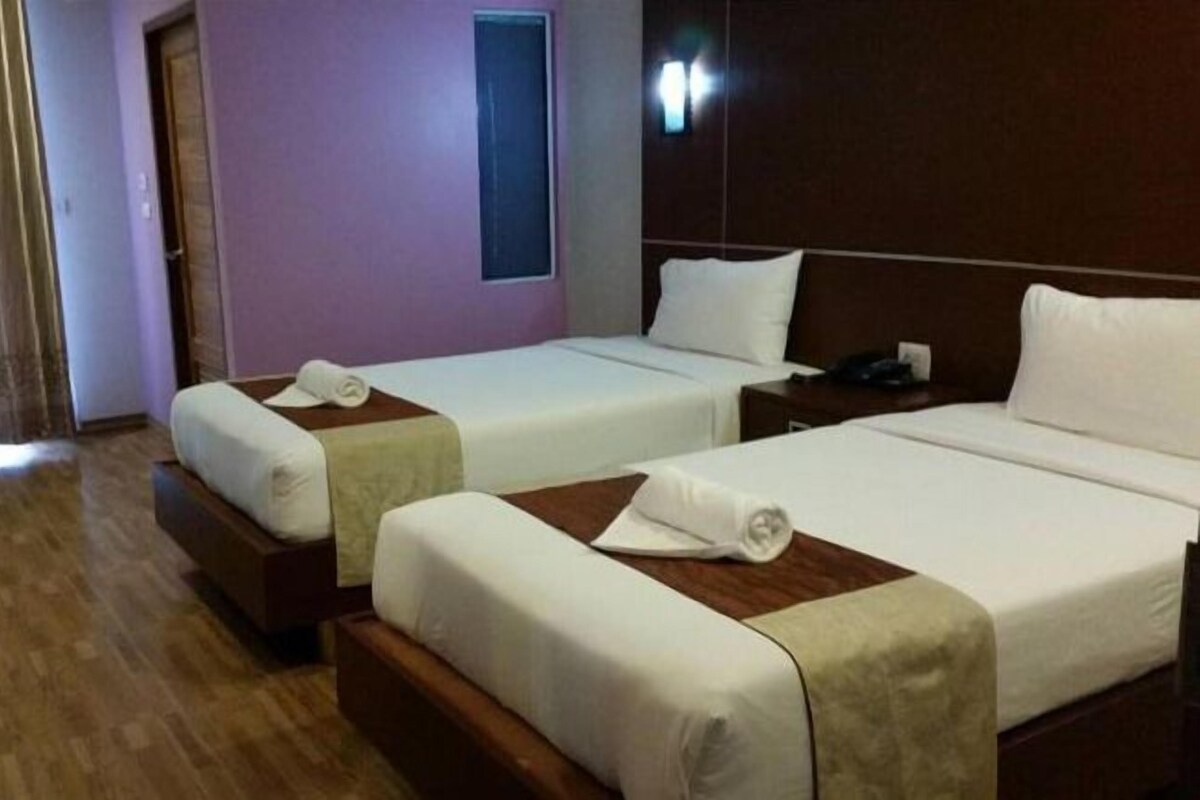 Una habitación de hotel con dos camas individuales, un cabecero de madera, una mesita de noche pequeña con una lámpara y una ventana con persianas.