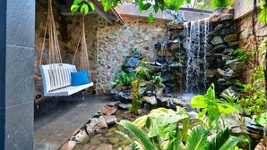 Un mur de pierres avec une petite cascade, une balançoire suspendue dotée d’un coussin bleu, et une variété de plantes tropicales.