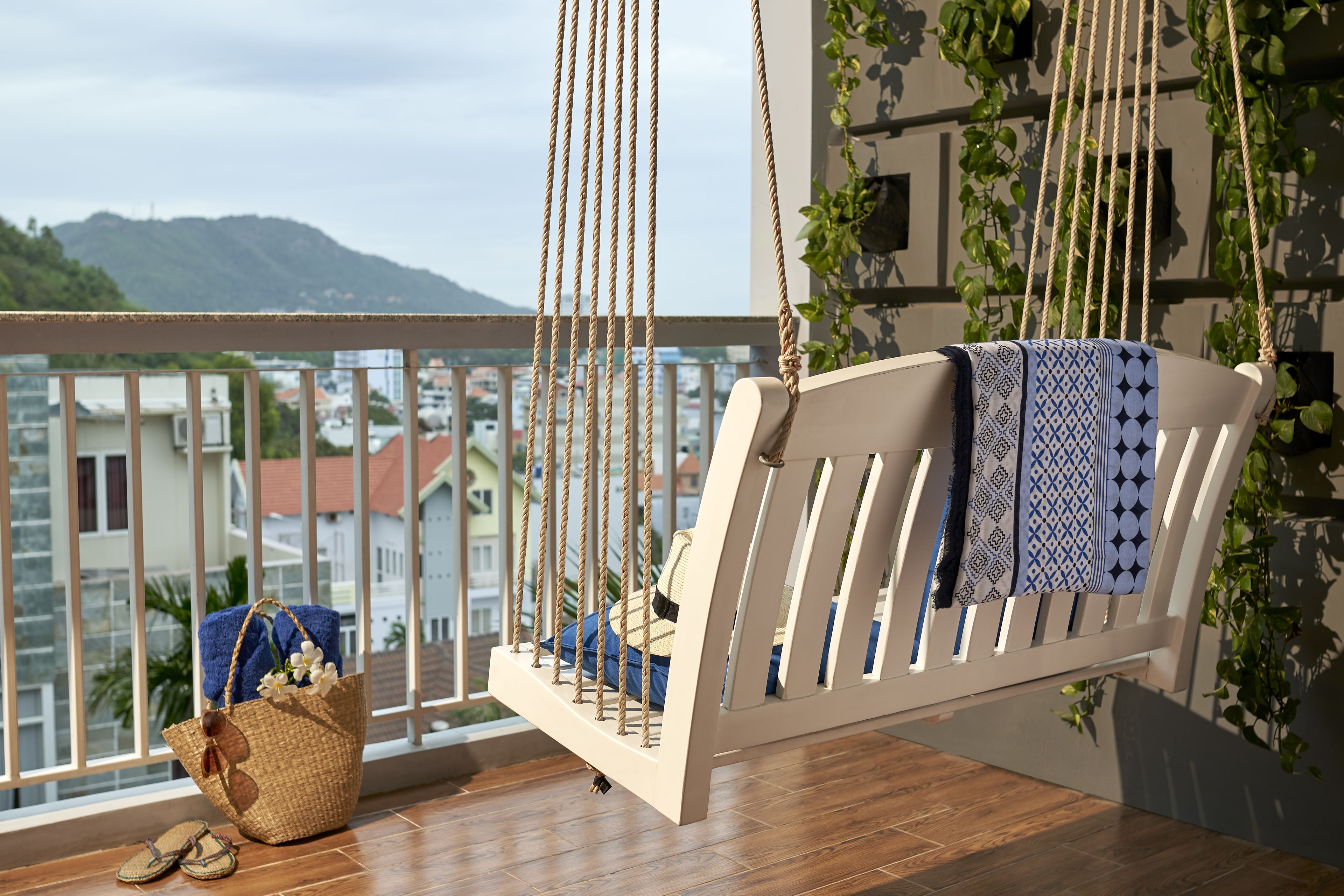 Une chaise suspendue en bois avec un coussin à motifs, un panier tressé et une paire de sandales, le tout sur un balcon offrant une vue sur des bâtiments et des collines.