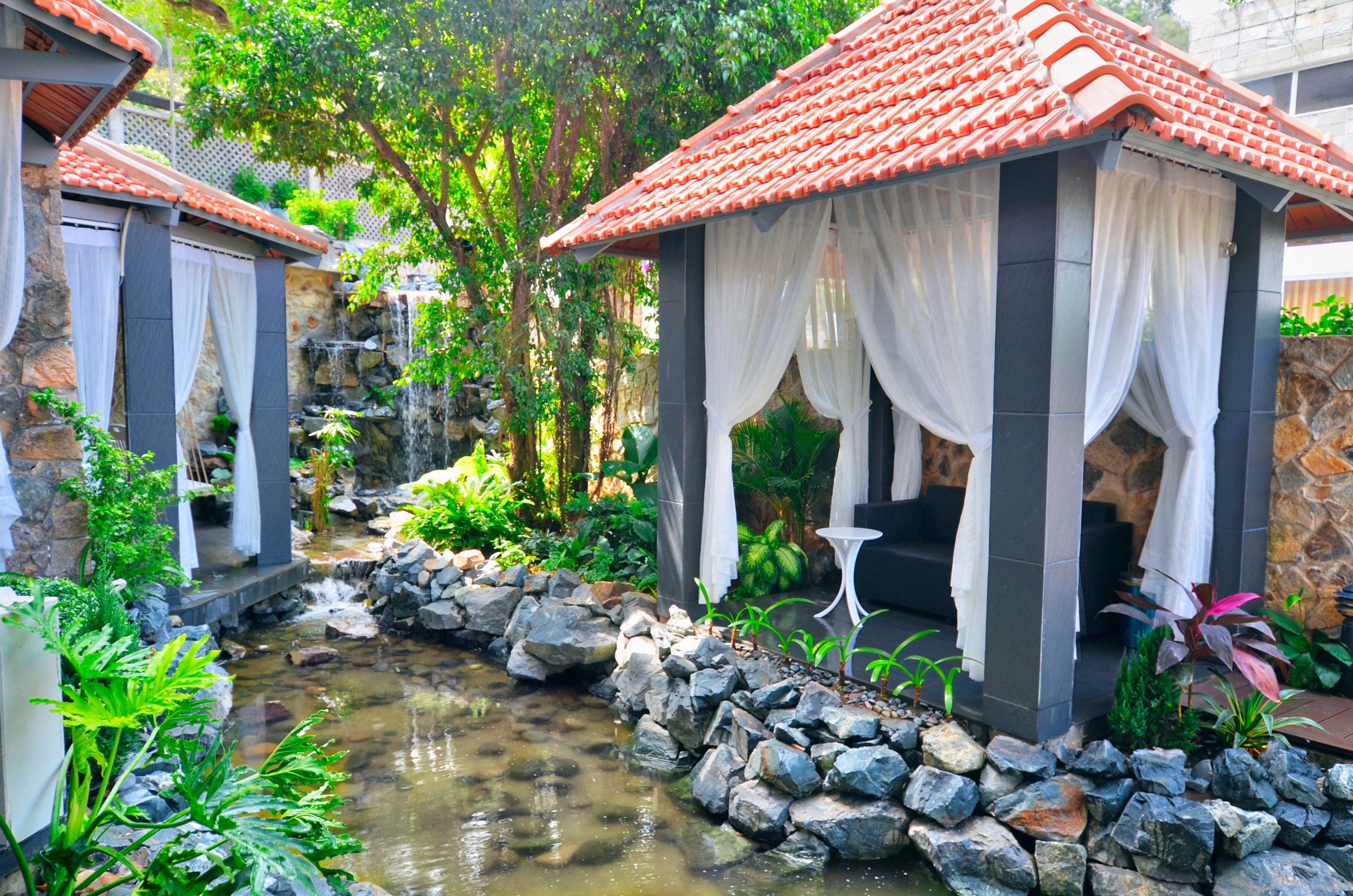 A serene garden with a small stream, stone walls, and a gazebo with white curtains.