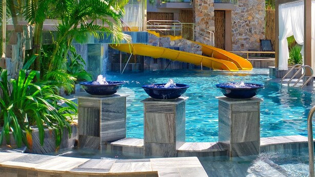 Une piscine avec un toboggan jaune, trois fontaines et des chaises longues.