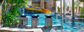 A pool with a yellow slide, three water fountains, and lounge chairs.