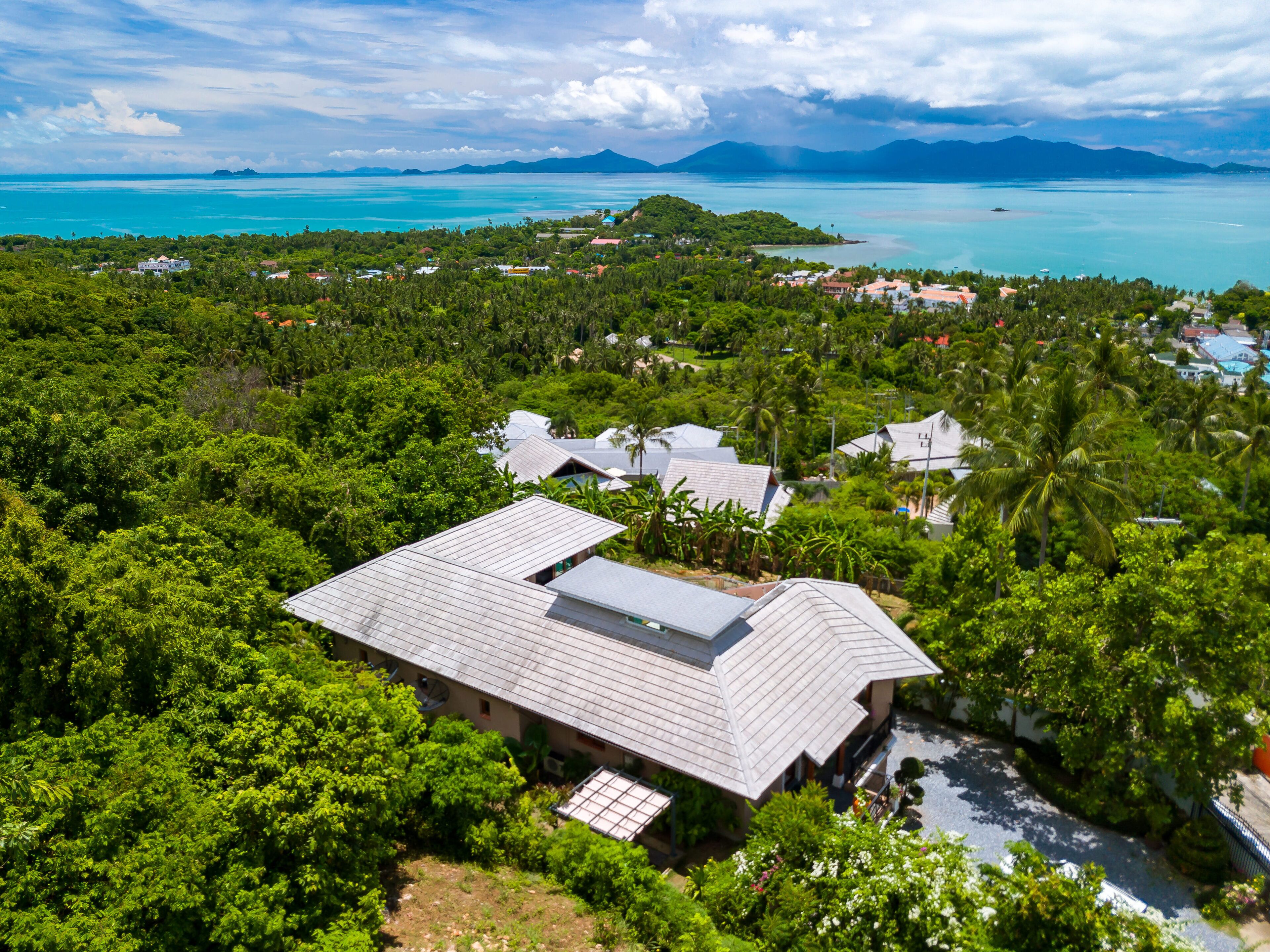 Une vue aérienne d’un complexe hôtelier tropical, composé de plusieurs bâtiments entourés d’une végétation luxuriante, offrant une vue dégagée sur l’océan et sur des montagnes lointaines.