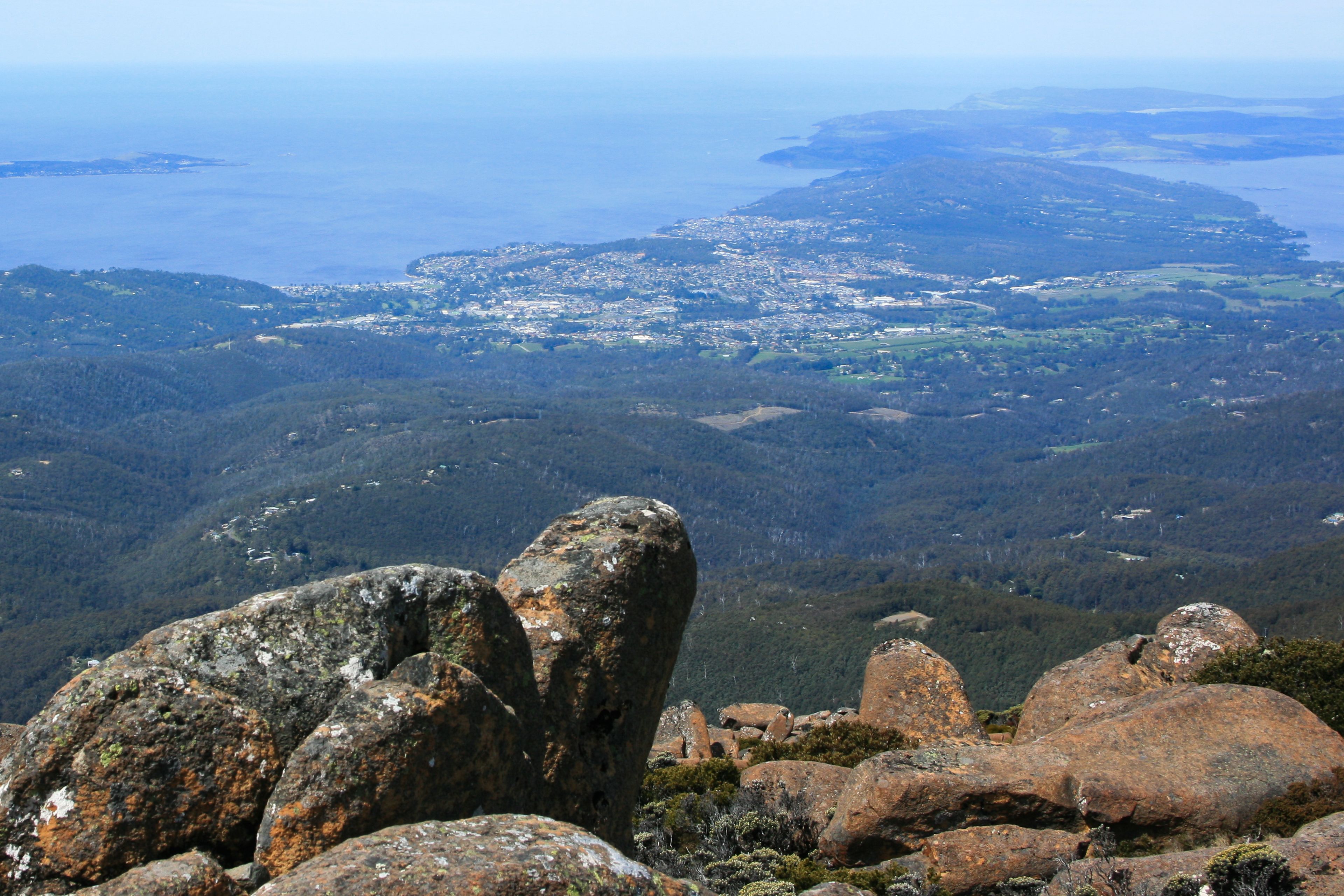 Un paysage montagneux, avec des formations rocheuses imposantes au premier plan et une vue lointaine d’une ville côtière.