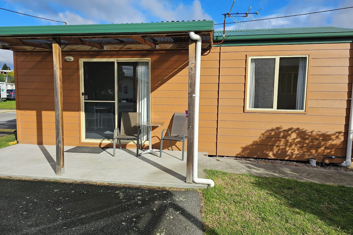 A small, single-story building with a green roof, a sliding glass door entrance, and a small patio area with a table and two chairs.