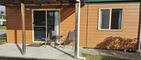 A small, single-story building with a green roof, a sliding glass door entrance, and a small patio area with a table and two chairs.