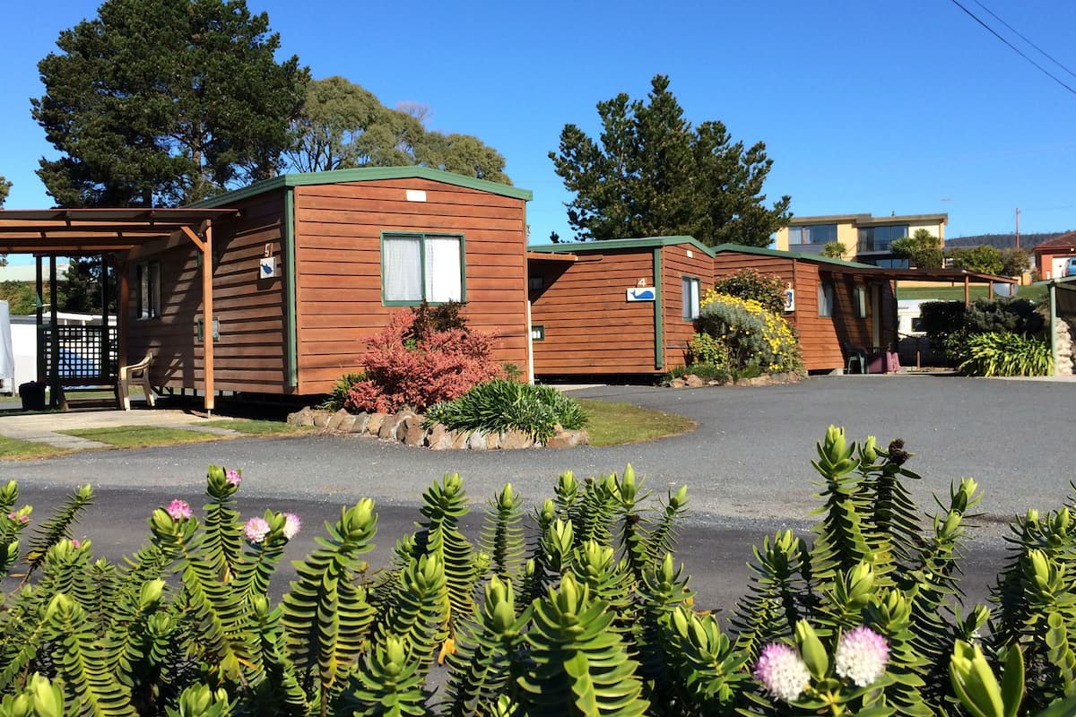 A row of wooden cabins with a covered porch, a small garden with pink flowers, and a clear blue sky.