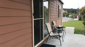 A covered patio with a table and chairs, a door, and a view of the street and greenery.