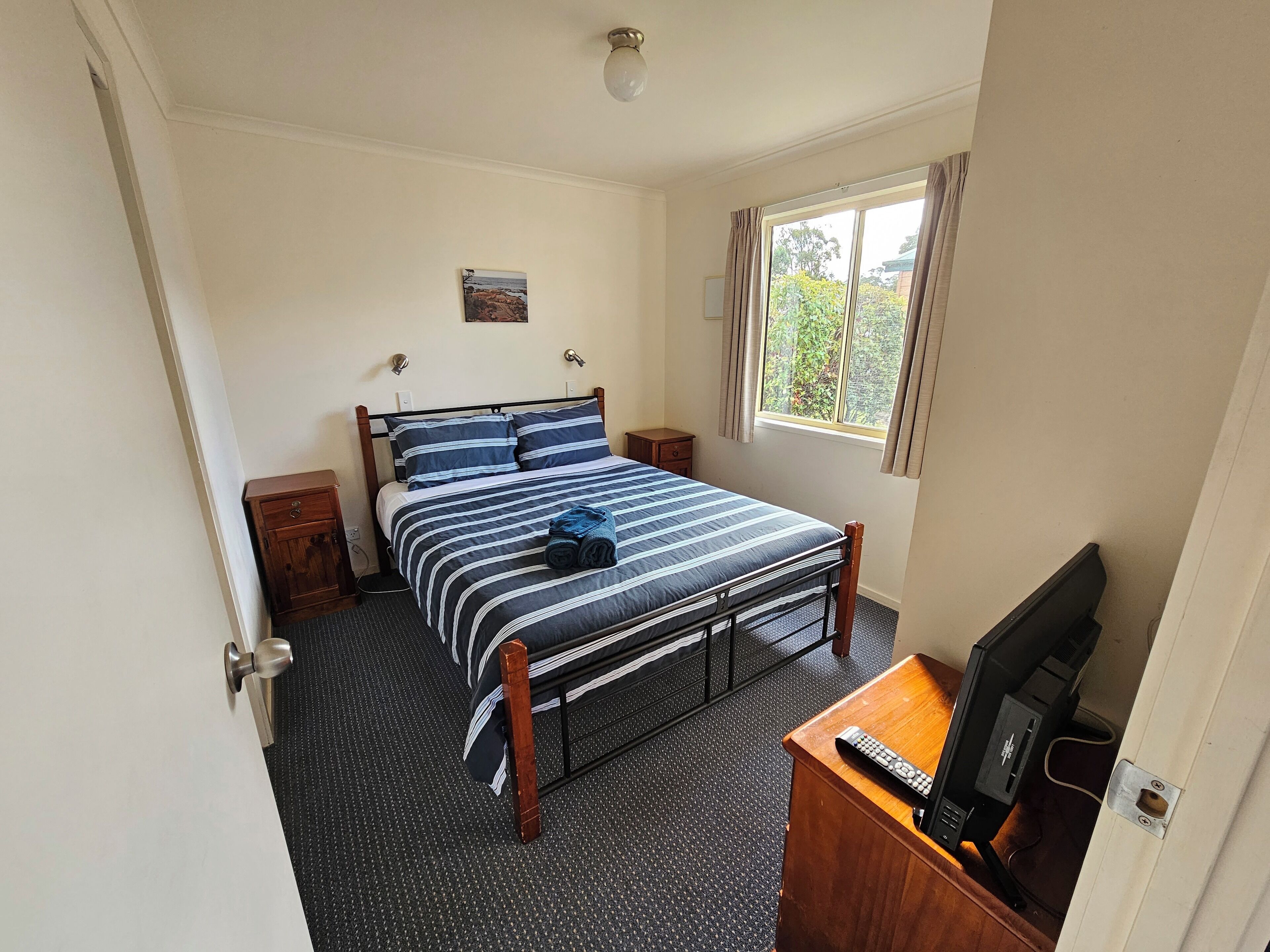 A bedroom with a bed, bedside tables, a desk with a television, a window with curtains, and a door.