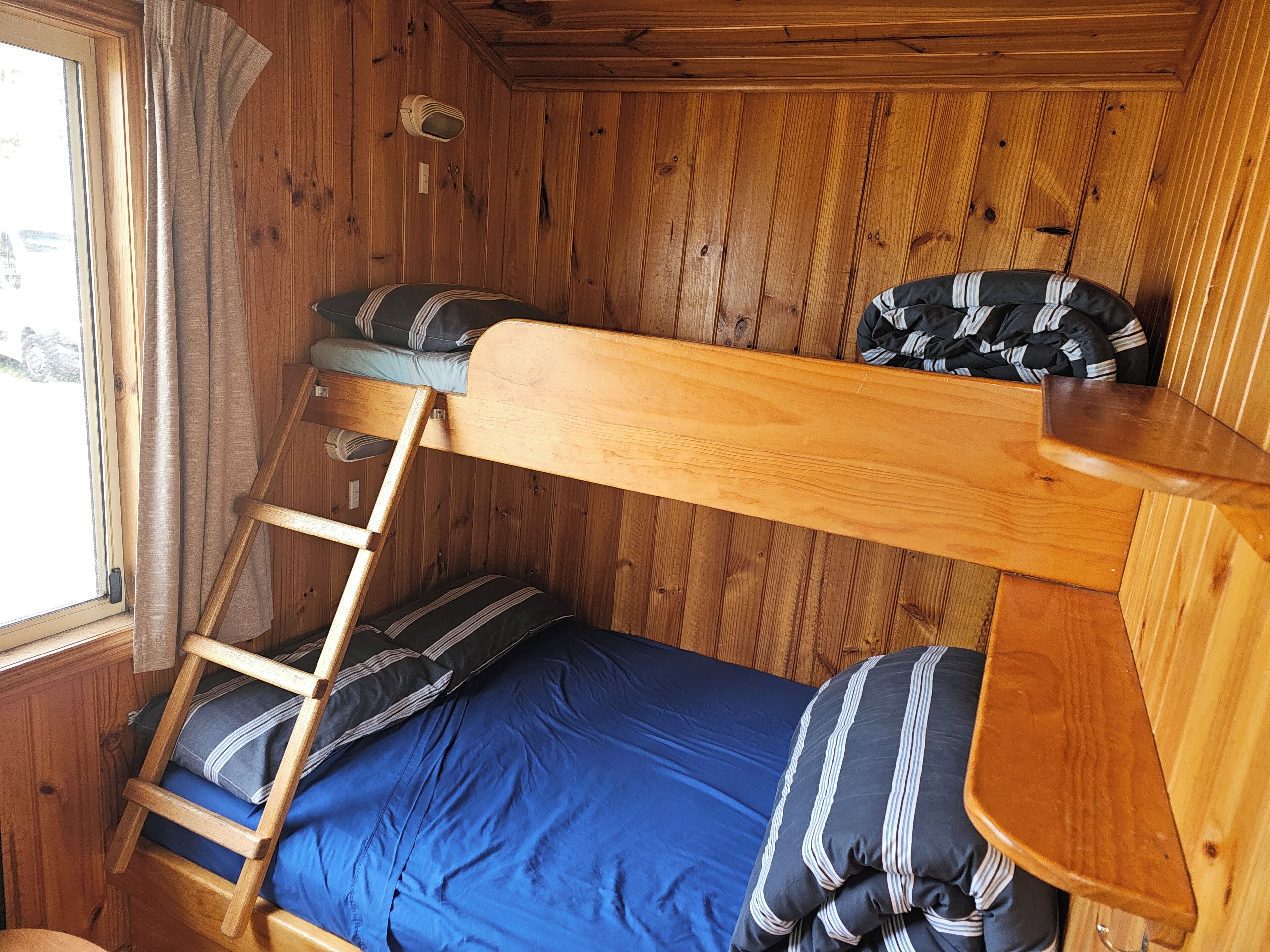 A bunk bed with a ladder, wooden walls, and a window with curtains.
