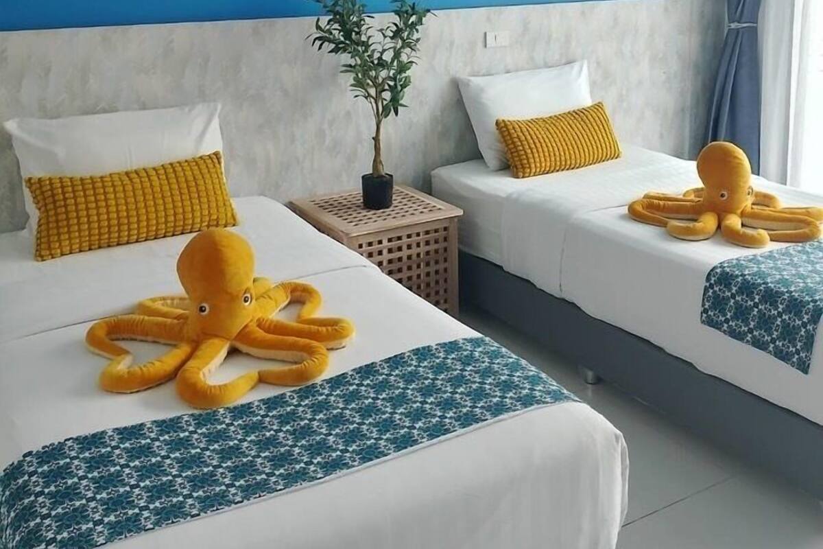 A hotel room with two beds, each with a white and blue patterned bedspread, a yellow pillow, and a large octopus plush toy on the bed. There are three framed artworks on the wall above the beds, a potted plant on a side table, and a window with curtains.