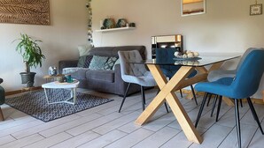 A modern living room with a glass dining table, blue chairs, a grey sofa, and a wooden ceiling.