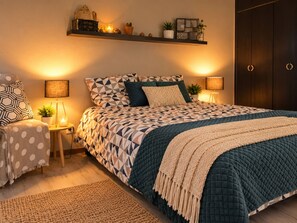 Un dormitorio con una cama, mesitas de noche, una lámpara, una repisa con objetos decorativos y una puerta.