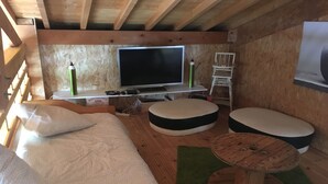 A cozy cabin interior with a bed, two round ottomans, a wooden coffee table, a television, and a small white chair. - Ideal house for 4 families with 2 children each  (Montgeron)