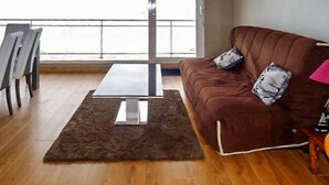 A living room with a brown sofa, a glass coffee table, a patterned rug, and a view through a large window. - Nice apartment with sea view a few meters away and balcony, close to shops. (Perros-Guirec)