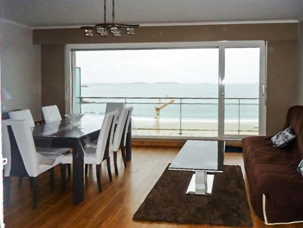 A modern living room with a dining area, a large sliding glass door leading to a balcony with a view of the ocean.