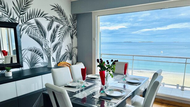 A dining area with a view of the sea, white chairs, a glass table, and a tropical leaf mural. - Nice apartment with sea view a few meters away and balcony, close to shops. (Perros-Guirec)