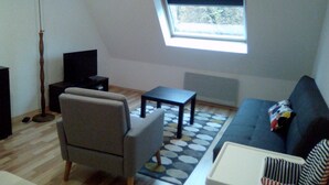 Un salon comprenant un canapé, un fauteuil, une petite table, un lampadaire et une fenêtre avec des stores.