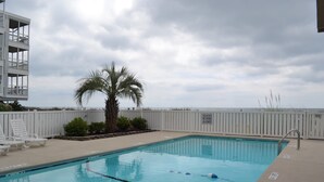 A swimming pool with lounge chairs and a palm tree, surrounded by a white fence and a building with balconies. - Comfy Direct Oceanfront Condo.  Our Happy Place can be Yours. Book a great deal! (North Myrtle Beach)