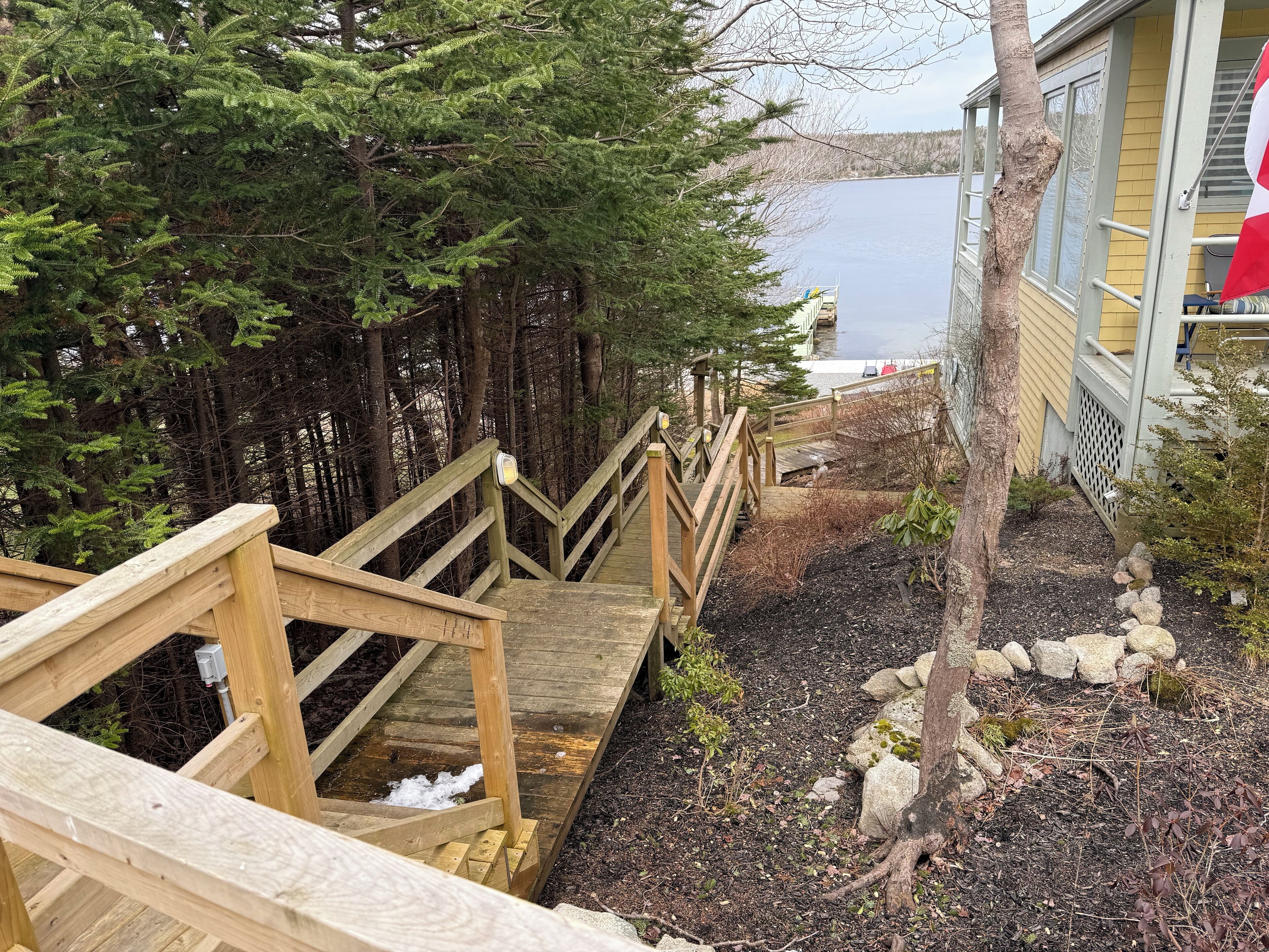 Un escalier en bois mène à une jetée surplombant une étendue d’eau, le tout étant entouré d’une forêt dense et d’un bâtiment à la façade jaune.