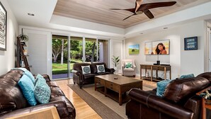 A spacious living room with leather sofas, a wooden coffee table, and a large window offering a view of a garden. - 🏖️Mauna Lani Townhome w/ Beach Club Access + A/C (Kamuela)