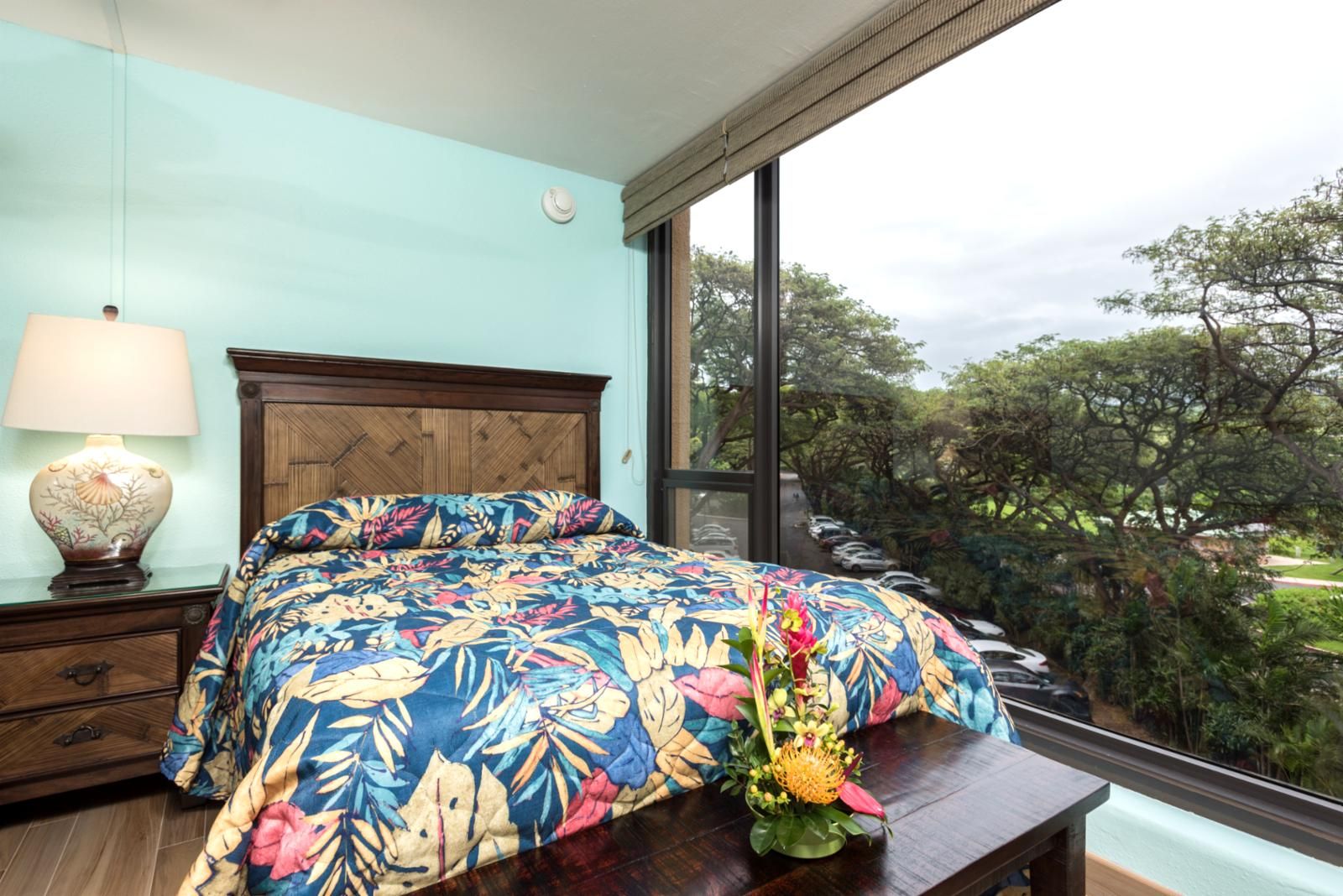 A bedroom with a large window, a wooden headboard, a floral bedspread, a bedside table with a lamp, and a view of trees and parked cars outside.