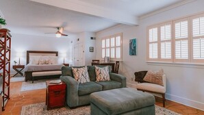 A spacious living room with a large sofa, a smaller sofa, and a bench. There is a wooden coffee table and a floor lamp. The room has a ceiling fan and a window with shutters. - Only 1 block to FRONT BEACH! Perfect location in Ocean Springs! The King Suite (Ocean Springs)