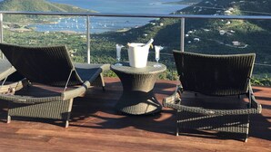 Een terras met rieten stoelen en een tafel, met uitzicht op een kustlandschap met heuvels en boten in de verte.