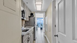 A narrow kitchen with white cabinets, a microwave, and a coffee maker. A door leads to a balcony with a view of the sky. - Scenic Views  Atlantica Resort 🏖️ Ocean Front🏖️ (Myrtle Beach)