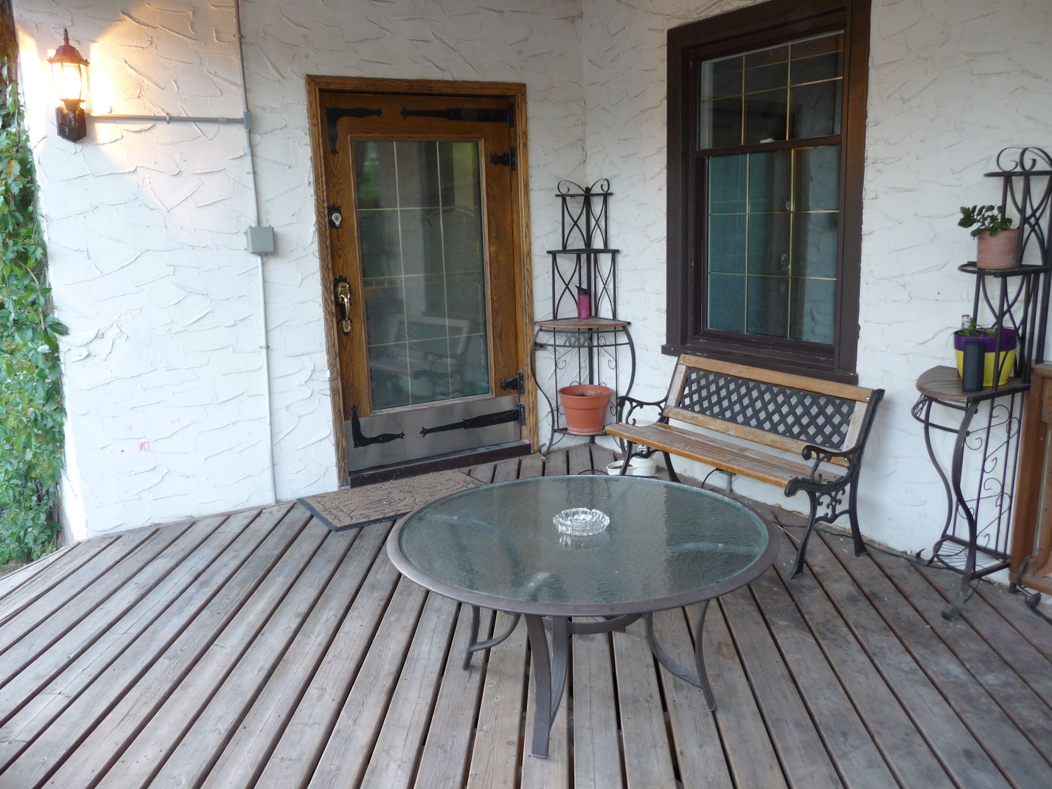 Una terraza de madera con una mesa de vidrio y bancos, una puerta con ventanal y macetas sobre soportes metálicos.
