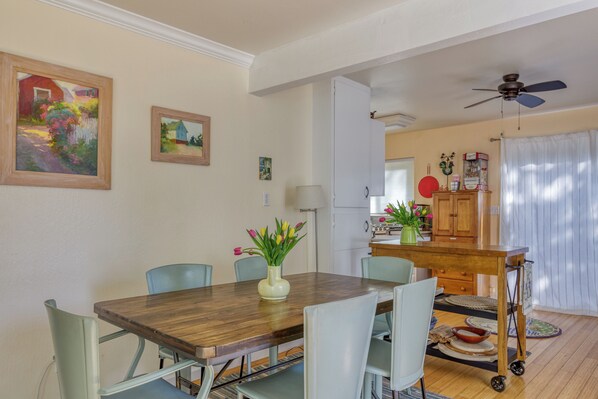 A dining area with a wooden table, light blue chairs, and a vase of flowers. There is a kitchen in the background with wooden cabinets and a ceiling fan.