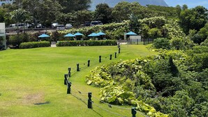 A lush, green lawn with a fenced area, surrounded by dense foliage and a mountain backdrop.