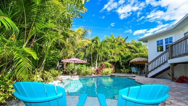 A pool area with two blue chairs, a covered seating area, and lush greenery. - Beach Time AMI! Pet Friendly Home with Pool, Hot Tub and Walk to Beach (Holmes Beach)