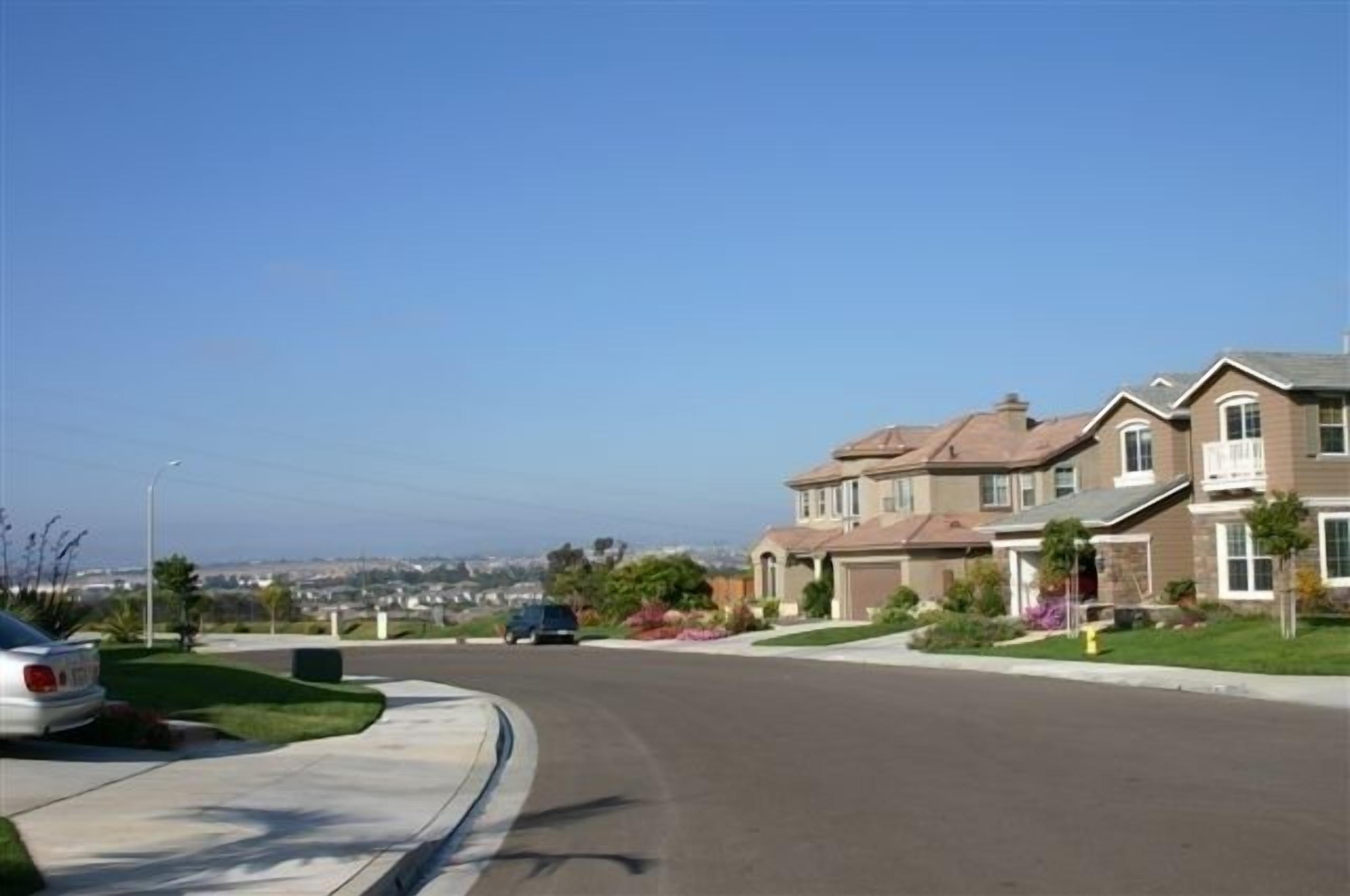 A residential street with two-story houses, a clear sky, and a distant cityscape.