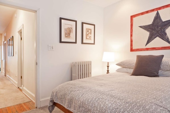 Une chambre à coucher avec un lit, un radiateur, une table de chevet, une lampe et deux tableaux encadrés accrochés au mur.