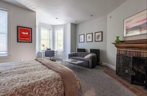 A modern bedroom with a large bed, a desk, a gray sofa, and a fireplace.