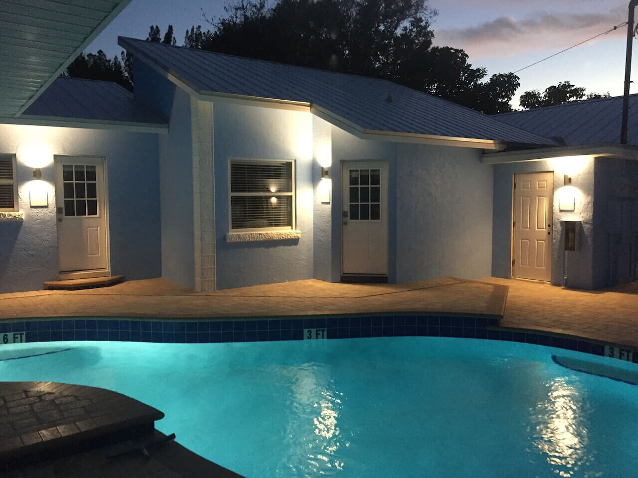 A residential swimming pool with a 6 ft and 3 ft depth marking, surrounded by a house with exterior lighting and a covered patio area.
