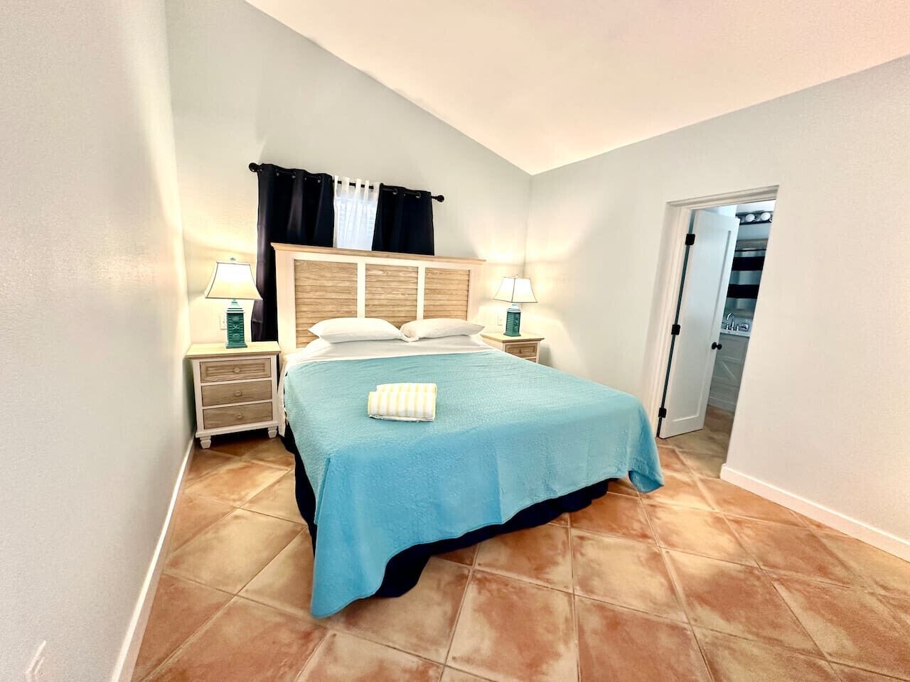 A bedroom with a bed, two bedside tables, a nightstand, a window with curtains, and a door leading to a bathroom.