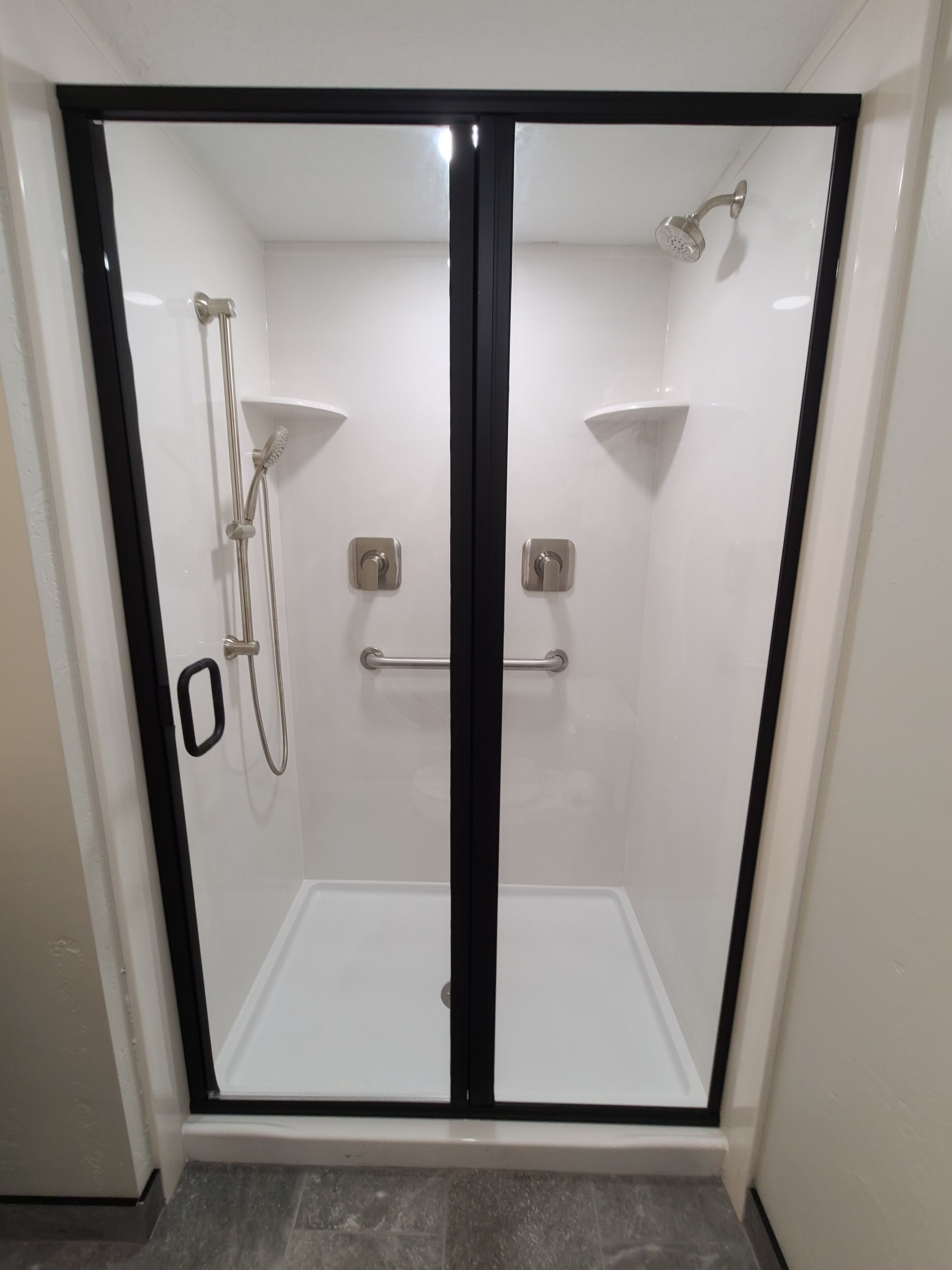 A glass-enclosed shower with a black frame, a handheld showerhead, a wall-mounted faucet, and a small shelf.
