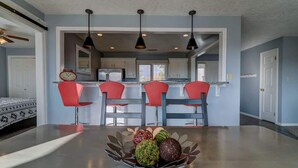 A modern kitchen with a dining area, featuring a central island with a fruit bowl, red chairs, and pendant lighting.